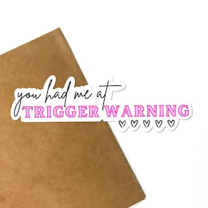 May include: A white sticker with black text that reads "you had me at TRIGGER WARNING" in pink letters.  Five small pink hearts are below the text.