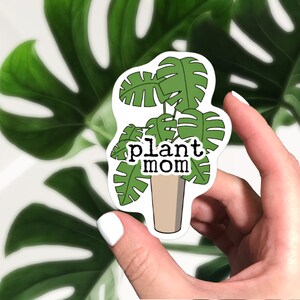 Puede incluir: Una pegatina blanca con una planta verde en una maceta marr&oacute;n. La planta tiene hojas grandes y el texto "plant mom" est&aacute; escrito en la pegatina.