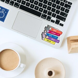 May include: A stack of colorful book stickers on a laptop. The books have titles like "Deal," "Mistake," "Score," "Goal," and "Legacy." A cup of coffee and a small dish are in the foreground.
