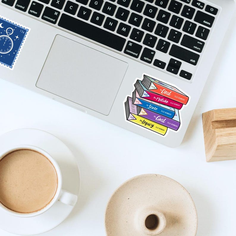 May include: A stack of colorful book stickers on a laptop. The books have titles like "Deal," "Mistake," "Score," "Goal," and "Legacy." A cup of coffee and a small dish are in the foreground.