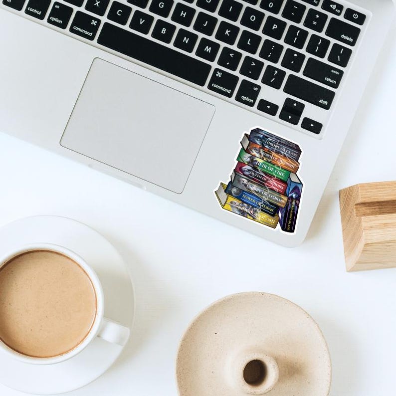 May include: A sticker featuring a stack of colorful books with titles visible, including "Throne of Glass" and "Heir of Fire." The sticker is placed on a laptop, with a coffee cup and a small dish in the background.