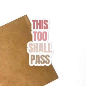 May include: A die-cut sticker featuring the text THIS TOO SHALL PASS in a stacked, block-letter format. The word THIS is in a dusty rose color, TOO is in a lighter pink, SHALL is in a pale beige, and PASS is in a medium brown tone.