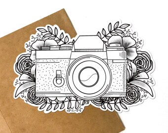 May include: A black and white line drawing of a vintage camera with a lens and a floral design around it.
