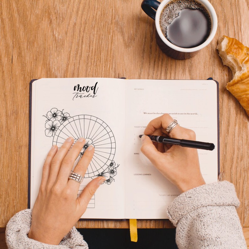 May include: A person is filling out a mood tracker journal with a black pen. The journal has a black and white floral design and the words "mood tracker" written in black cursive. The journal is open to a page with a circular mood tracker, a section for "today's tasks", "wins", and "lessons learned".