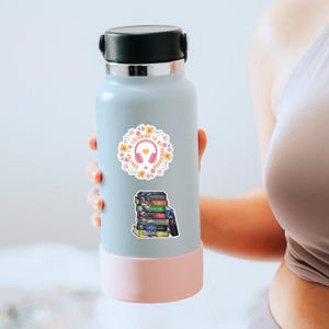 May include: A light blue water bottle with a black cap and a pink base. The bottle has two stickers: one with headphones and the text "I'm listening to my audiobook" and another with a stack of books. The bottle is held by a person.
