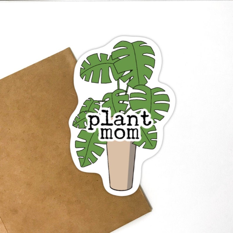 Puede incluir: Una pegatina con una planta monstera verde en una maceta beige con el texto "plant mom" en negro.