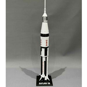 Può includere: Un modello di razzo Saturn 1B, prevalentemente bianco con dettagli neri e argento. La parola "USA" è stampata in rosso sul lato. Il razzo è posizionato su una base nera con la scritta "SATURN 1B".