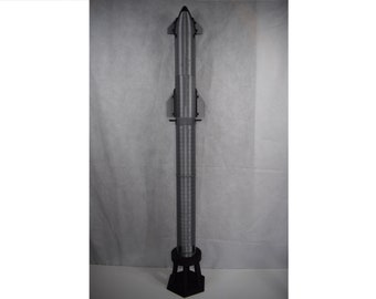 Può includere: Un modello di razzo grigio e nero su un supporto nero. Il razzo è alto e sottile con un cono di naso appuntito e una serie di alette alla base.