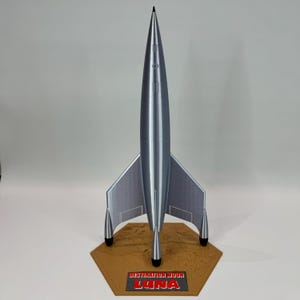 May include: A silver model rocket with a pointed nose cone and three fins. The rocket is standing on a hexagonal base with the words "Destination Moon LUNA" printed on it.