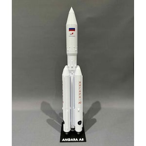 May include: A white model rocket with the text "ANGARA A5" on a black base. The rocket has a pointed nose cone and features the Russian flag and the logo of Roscosmos. The rocket is a detailed replica of a space launch vehicle.