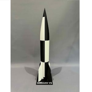 May include: A black and white model rocket with a pointed top, standing on a black base. The rocket has a checkerboard pattern and the text "GERMAN V2" is printed on the base.