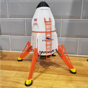 May include: A white and orange model of a SpaceX Starship with a ladder on the side and the text "SpaceX" printed on the side. The model has three orange legs with black feet.
