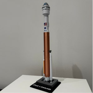 Op de afbeelding: Een model van de Atlas V Starliner-raket, met een koperkleurig middengedeelte, witte bovenste trappen en een zwarte basis. Het model toont het NASA-logo en de Amerikaanse vlag. De basis is gemarkeerd met "ATLAS V STARLINER".