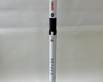 Può includere: Un modello di razzo bianco e nero con le parole "SpaceX" e "NASA" stampate sul lato. Il razzo è su un supporto nero con tre gambe.