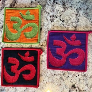 May include: Three colorful pot holders with the symbol Om embroidered on them. The pot holders are orange, purple, and black with red embroidery.