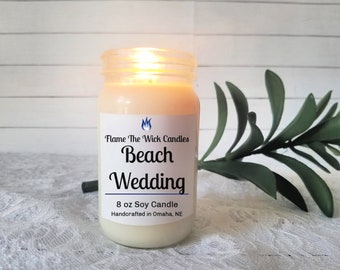 May include: A lit soy candle in a clear jar with the text "Beach Wedding". The candle is 8 oz and handcrafted in Omaha, NE. A green plant is next to the candle.