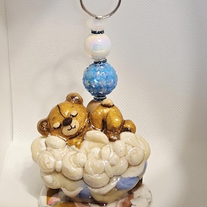 May include: A handmade keychain featuring a sleeping teddy bear on a cloud. The bear is gold and brown, resting on a white cloud base. The keychain has a silver ring and decorative beads in white and blue.