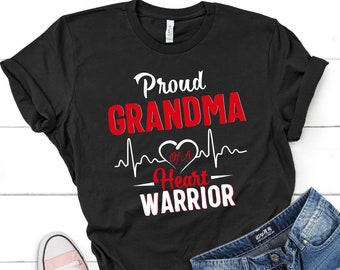May include: A black short-sleeved t-shirt featuring the white and red text Proud GRANDMA OF A Heart WARRIOR with a white heartbeat line graphic forming a heart shape in the center. The shirt is displayed alongside blue denim shorts and a pink sneaker.