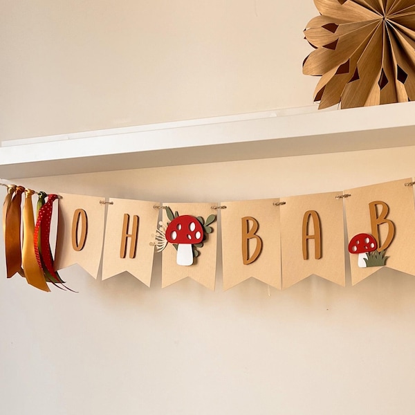 May include: A banner with the letters "OH BAB" on kraft paper pennants. The banner is decorated with wooden letters, red and white mushroom cutouts, and green foliage. Colorful ribbons hang from the left side. A decorative paper fan is in the background.