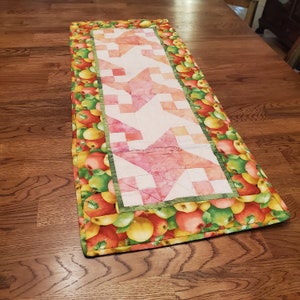 May include: Quilted table runner featuring a central star pattern in shades of pink and peach. The runner is bordered with a vibrant apple print in red, green, and yellow, adding a touch of rustic charm to any table setting.