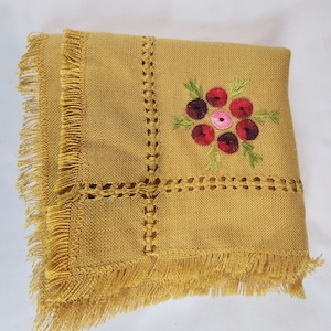 May include: Mustard-colored square tablecloth with a fringed edge and embroidered floral design. The embroidery features red, pink, and purple flowers with green leaves. Decorative stitching frames the floral design.
