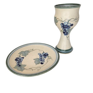 May include: Hand-painted ceramic plate and goblet set. The off-white pieces feature blue grape clusters and green leaves, with blue and green trim. The goblet is approximately 7 inches tall.