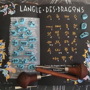 Peut inclure: Un carnet noir avec le titre "LANGUE DES DRAGONS" écrit en blanc. Le carnet contient un alphabet manuscrit avec des symboles en or et en bleu. Il y a aussi des tampons en caoutchouc bleu avec des symboles dessus. Deux outils en bois sont sur la page.
