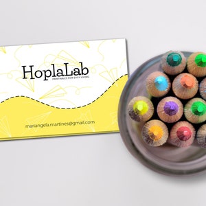 May include: A yellow business card with the text "HoplaLab Printables for Easy Living" and the email address "mariangela.martines@gmail.com". A close-up of a set of colored pencils in a brown container.