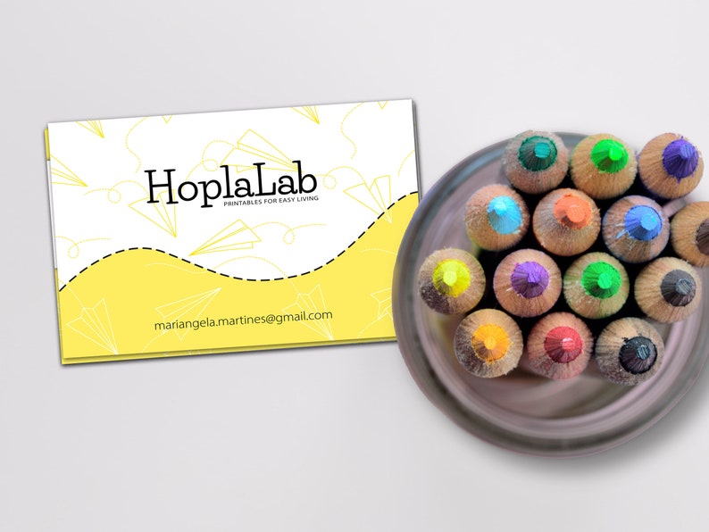 May include: A yellow business card with the text "HoplaLab Printables for Easy Living" and the email address "mariangela.martines@gmail.com". A close-up of a set of colored pencils in a brown container.