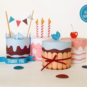 May include: Three printable papercraft birthday cake boxes. The cakes are decorated with frosting, sprinkles, candles, and a cherry. The boxes have a "Thank You" banner.