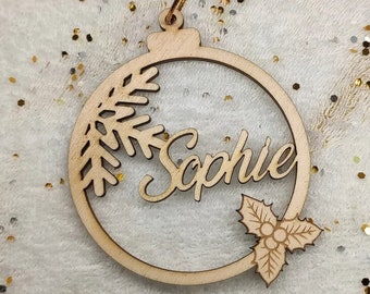 May include: A wooden Christmas bauble shaped like a circle with a cutout design of a pine branch, the name "Sophie" and a holly sprig. The bauble is hanging from a gold string.