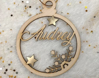 May include: A wooden Christmas bauble with the name "Audrey" cut out in the centre. The bauble is decorated with gold stars and a snowflake.