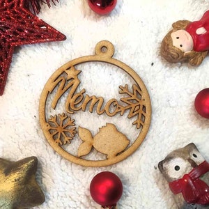 May include: Wooden Christmas ornament with the word "Mémos" cut out, along with snowflakes, a star, and a fish. The ornament is surrounded by red Christmas decorations, including a star, ornaments, and a figurine.