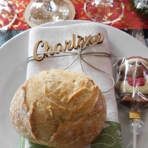 Op de afbeelding: Een witte linnen servet met een houten naamschildje waarop "Charlene" staat, vastgebonden met touw. Een rond broodje ligt op de servet naast een chocolade berenlolly.