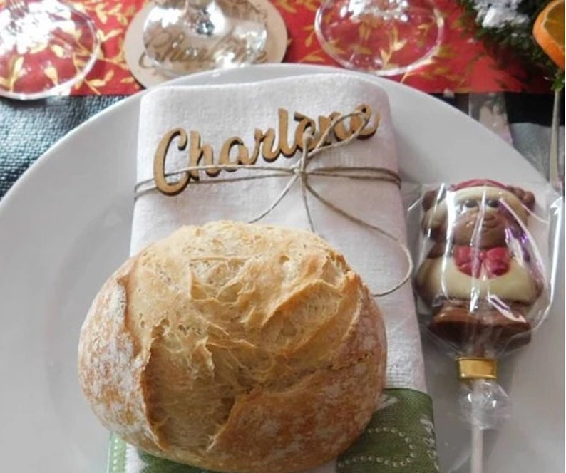 Op de afbeelding: Een witte linnen servet met een houten naamschildje waarop "Charlene" staat, vastgebonden met touw. Een rond broodje ligt op de servet naast een chocolade berenlolly.
