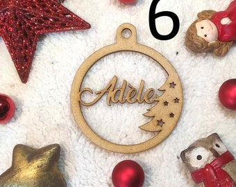 May include: A wooden Christmas bauble with the name "Adele" cut out, framed by a circle with a tree and stars. The number "6" is in the upper right corner. Red and gold Christmas decorations surround the bauble.