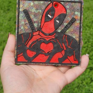 May include: A red and black Deadpool character figurine with a heart-shaped hand gesture. The figurine is made of resin and has a glittery background.