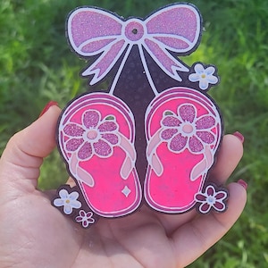 May include: A pink glittery bow tops a black silhouette of two pink flip-flops with flower accents. The flip-flops have pink straps and flower details. Small white and pink flowers are scattered around the base of the design.