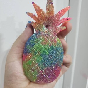 May include: A rainbow glitter pineapple shaped air freshener. The pineapple is made of a clear resin with a glittery finish. The pineapple has a hole in the top for hanging.