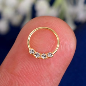 Può includere: Un anello per il naso color oro con cinque gemme chiare e scintillanti disposte in fila. L'anello è circolare e sembra essere fatto di metallo lucido. Le gemme sono piccole e ben distanziate.
