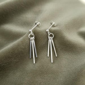 May include: A pair of silver dangle earrings. Each earring features a post, a small ball, a ring, and three vertical bars of varying lengths. The earrings are displayed on a textured, olive-green surface.