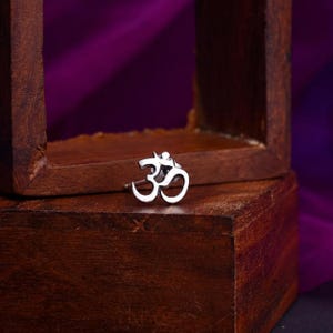 May include: A small silver-toned metal nose stud featuring the Om symbol. The jewelry piece has a polished, reflective surface and a curved post extending from the back, designed for piercing placement with a clear, recognizable shape.