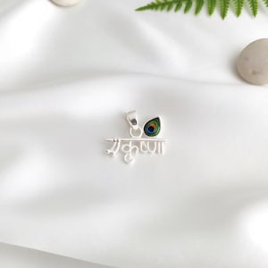 May include: A silver pendant with the word "Krishna" in a decorative script. The pendant has a small, colourful peacock feather design. The pendant is shown on a white, textured surface.