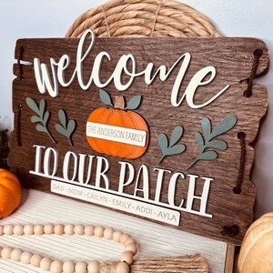 Puede incluir: Un letrero de madera rústico con la inscripción "Bienvenido a nuestro parche" y "The Anderson Family". El letrero está decorado con una calabaza y hojas.