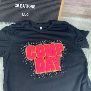May include: A black short-sleeved t-shirt featuring the text COMP DAY in bright pink letters with an orange outline. The text is surrounded by a scattering of small, shimmering rhinestones. A black letter board displays WALNUT ACRES CREATIONS LLC.