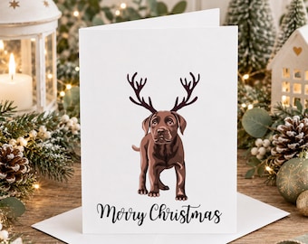 May include: A white greeting card featuring a brown Labrador puppy wearing dark brown reindeer antlers on its head. Below the puppy, the text Merry Christmas is printed in a black cursive font. The card is folded and stands upright.