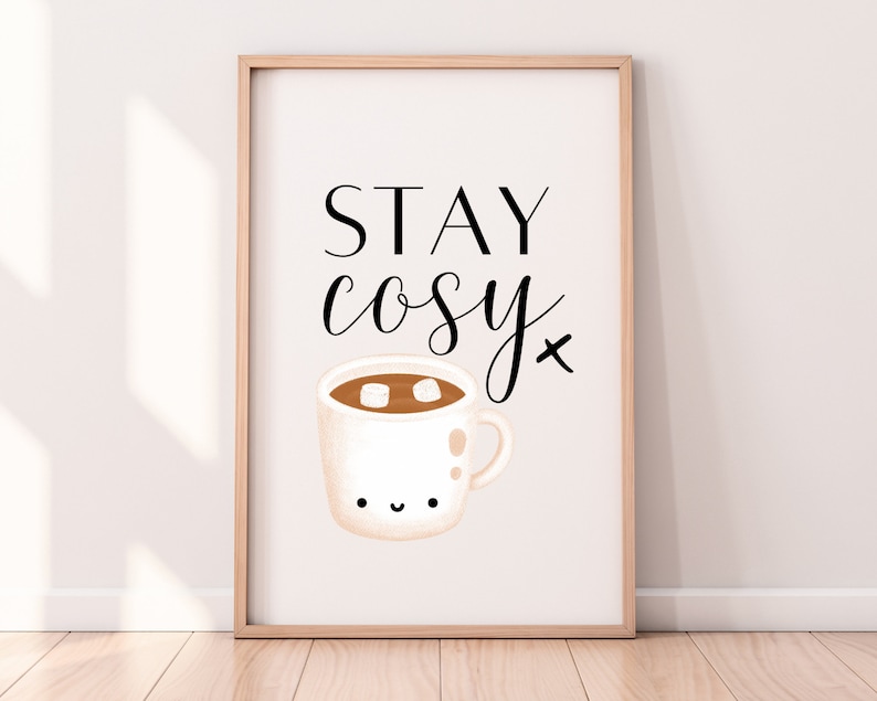 Puede incluir: Una impresi&oacute;n enmarcada en blanco con el texto "STAY cosy" en fuente cursiva negra. Debajo del texto hay una ilustraci&oacute;n de dibujos animados de una taza blanca con un l&iacute;quido marr&oacute;n y malvaviscos blancos en su interior.