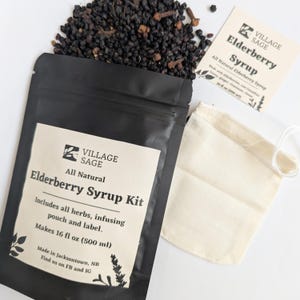 Könnte beinhalten: Ein schwarzer Beutel mit der Aufschrift Village Sage All Natural Elderberry Syrup Kit, dazu ein Haufen getrockneter dunkler Beeren und Nelken, ein kleiner beiger Stoffbeutel zum Aufbrühen und ein Etikett für 500 ml Sirup.