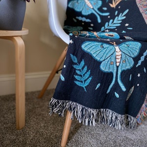 May include: A black woven throw blanket with a fringe border features a repeating pattern of blue and green moths and leaves on a dark background.