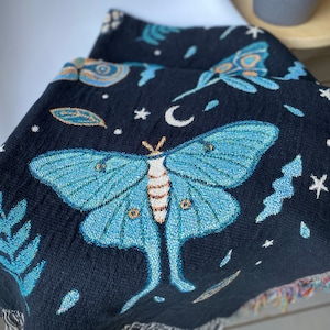 May include: A black woven tapestry throw blanket with a blue and green luna moth design. The blanket features a crescent moon, stars, and leaves.
